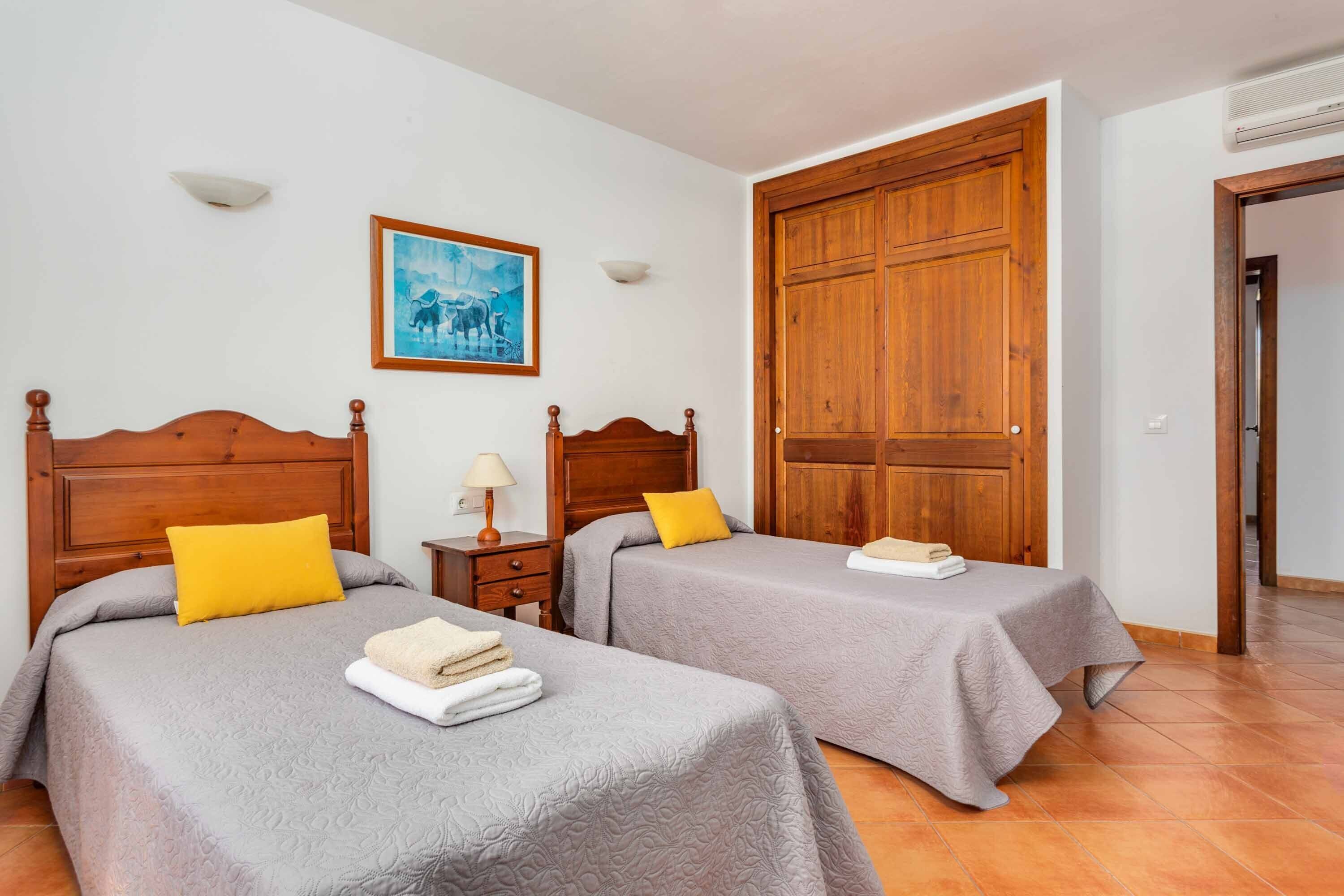 Ein Hotelzimmer mit zwei Betten, jeweils mit einem Holz-Kopfteil und einer grauen Tagesdecke. Darüber hängt ein Gemälde an der Wand, es gibt einen Holzschrank und einen kleinen Nachttisch mit einer Lampe.