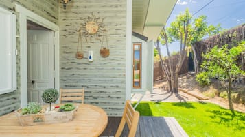 Een houten terras met een tafel en stoelen, een kleine tuin en een decoratieve muur met een zon en hangende decoraties.