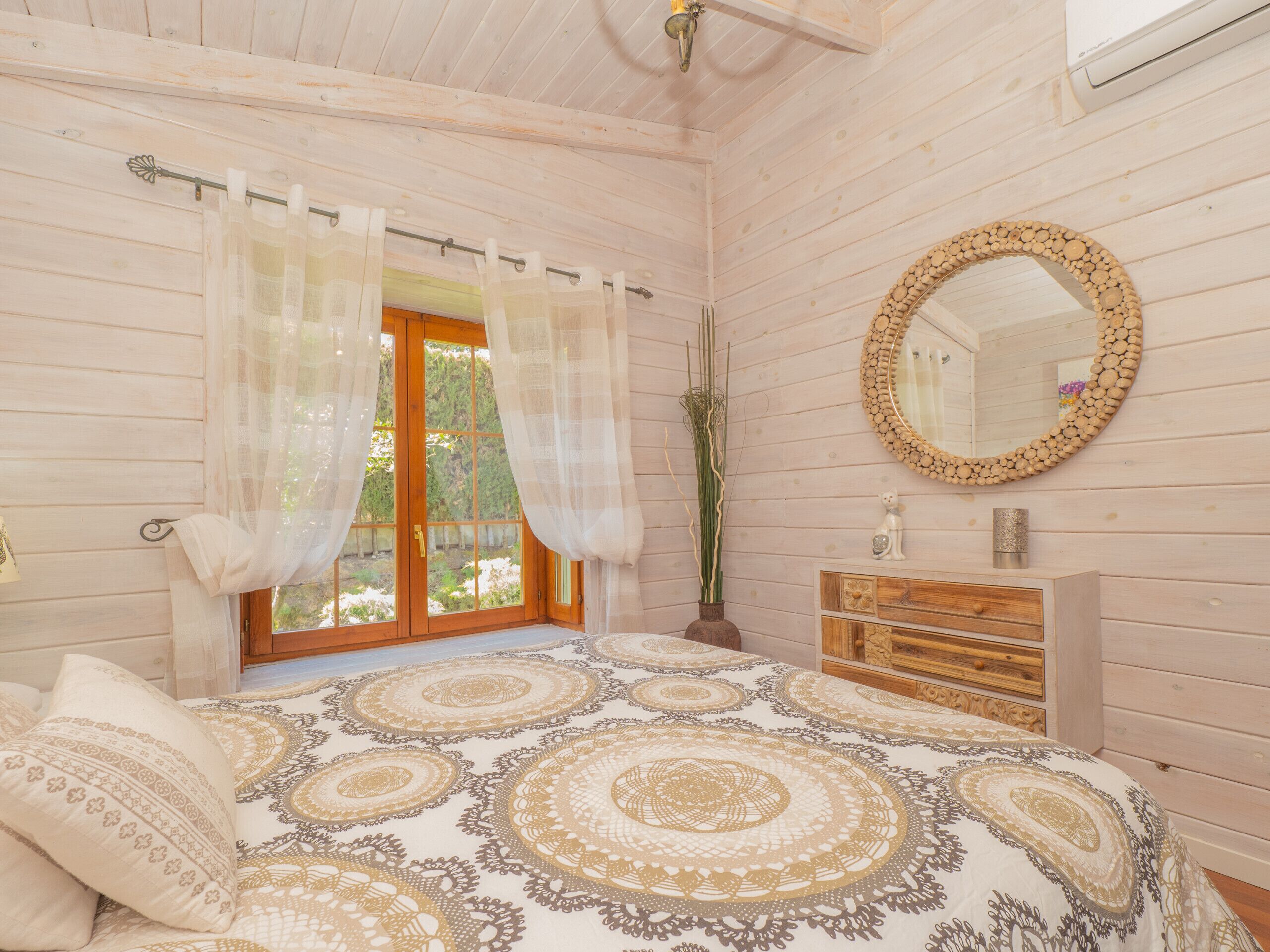 Een slaapkamer met een groot bed, een houten dressoir, een spiegel en een raam met gordijnen.