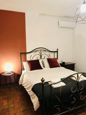 A neatly made bed with a black metal frame and white bedding, a small bedside table with a lamp, and a wall-mounted air conditioner. - Carabollace Estate - Mistral (Sciacca)