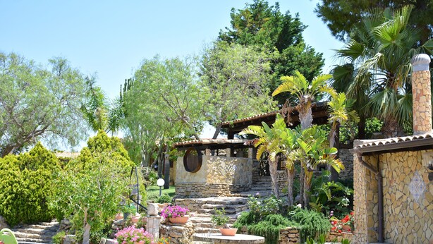 A stone building with a tiled roof, surrounded by lush greenery and various plants. - Carabollace Estate - Mistral (Sciacca)