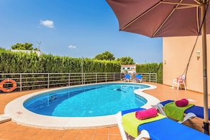 A swimming pool with lounge chairs and umbrellas, surrounded by a hedge and a building.