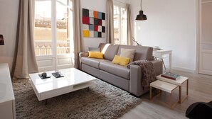 En moderne stue med sofa, sofabord og et lille sidebord. Et farverigt abstrakt kunstværk pryder væggen, og store vinduer giver udsigt til det fri.