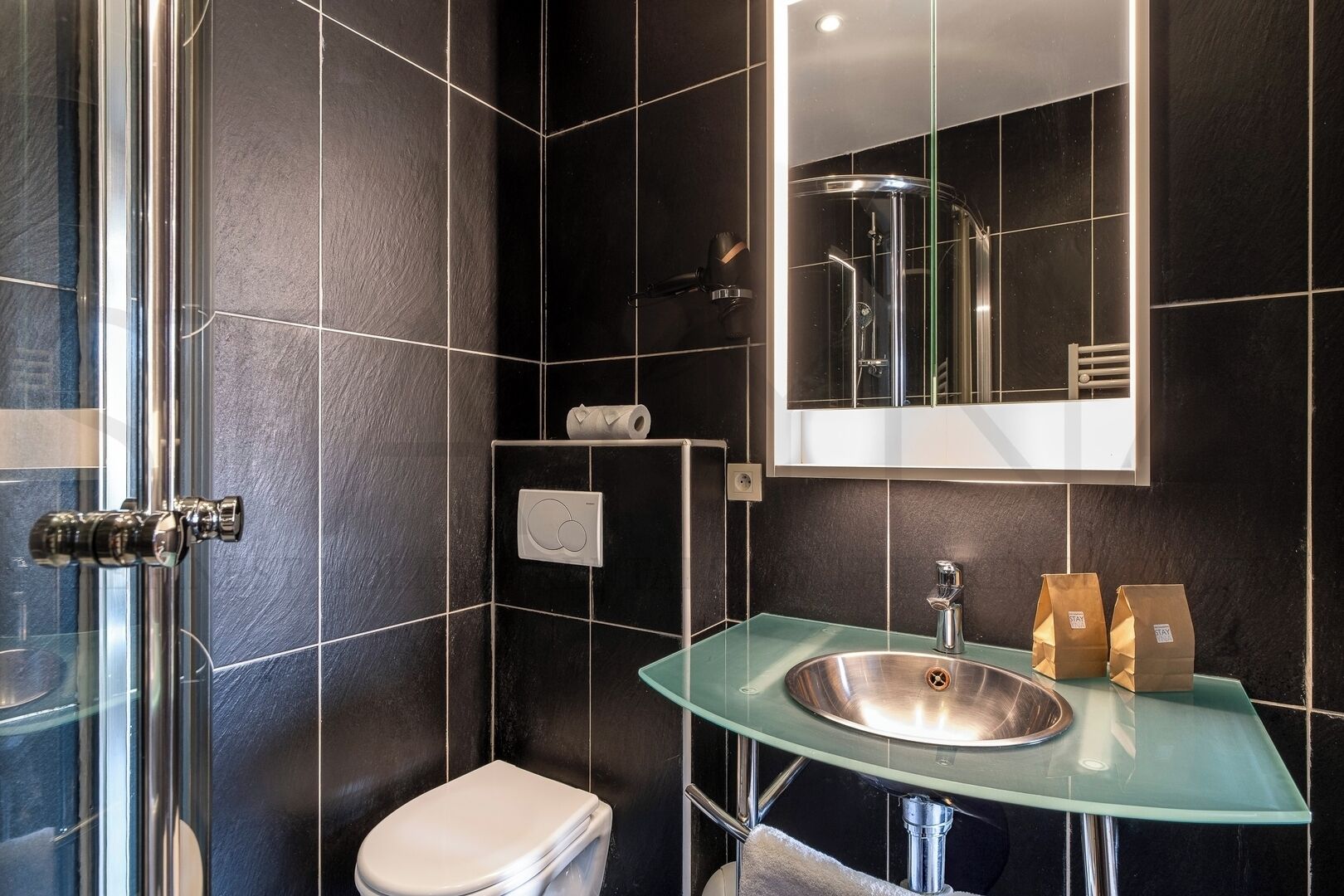 A modern bathroom with a glass sink, a toilet, and a shower. There are two brown paper bags on the sink.