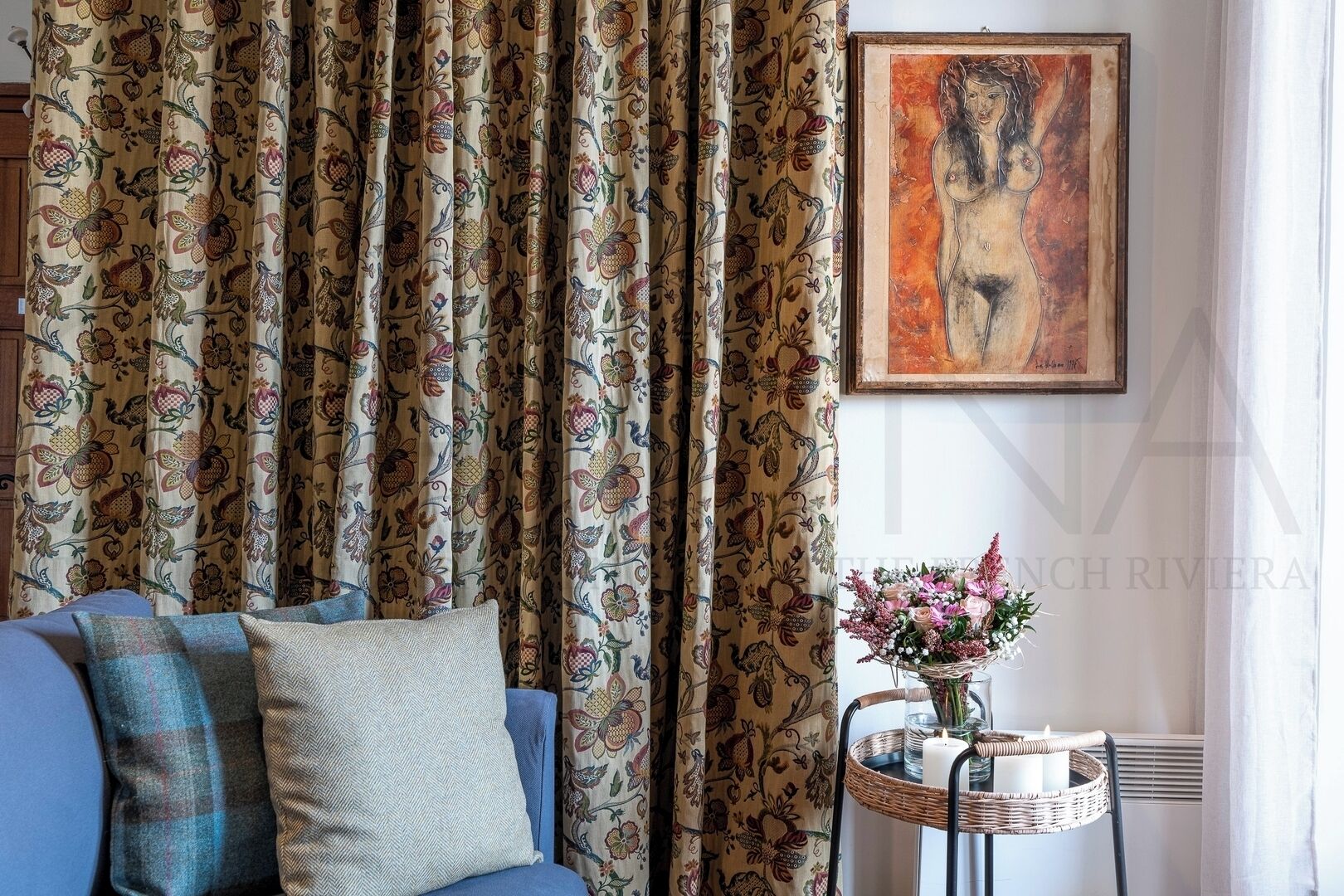 Un salon avec un canapé, un rideau à motifs, un tableau accroché au mur et une petite table sur laquelle se trouvent une bougie et des fleurs.