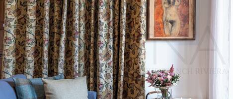 Un salon avec un canapé, un rideau à motifs, un tableau accroché au mur et une petite table sur laquelle se trouvent une bougie et des fleurs.
