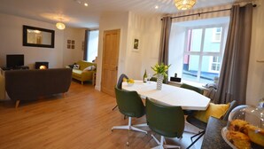 A modern living room with a dining area, wooden flooring, a fireplace, and a large window with curtains. - Morning Side 4 bedroom house VRBO (Dingle Peninsula)
