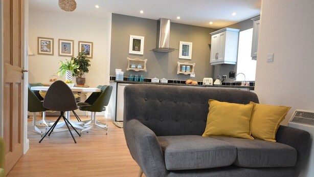 A modern living room with a grey sofa, a dining area with a table and chairs, and a kitchen with cabinets and a stove. - Morning Side 4 bedroom house VRBO (Dingle Peninsula)