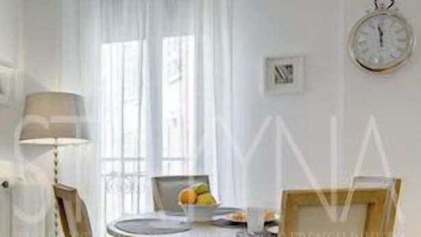 A dining area with a round table, two chairs, a clock on the wall, and a window with sheer curtains. - Minuet - 1 bedroom apartment (Nice)