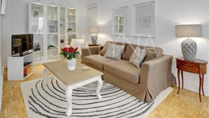 A modern living room with a beige sofa, a coffee table, and a TV stand. There are white shelves with decorative items and a lamp on a side table. - Minuet - 1 bedroom apartment (Nice)