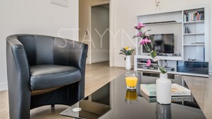 A modern living room with a black leather sofa, a glass coffee table, a white vase with flowers, and a glass of orange juice. There is a bookshelf with books and a flat-screen TV. - Coco 2 bedroom apartment VRBO (Nice)