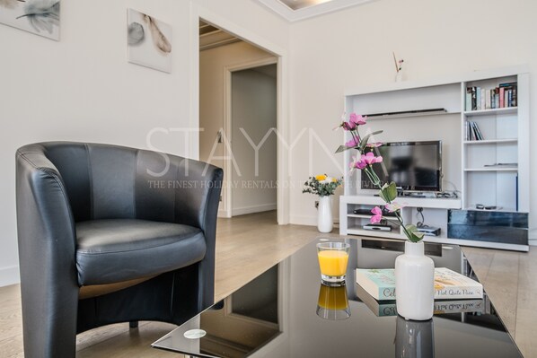 A modern living room with a black leather sofa, a glass coffee table, a white vase with flowers, and a glass of orange juice. There is a bookshelf with books and a flat-screen TV. - Coco 2 bedroom apartment VRBO (Nice)