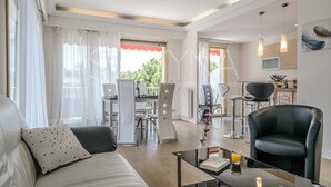 A modern living room with a sofa, a coffee table, and a black leather chair. There is a dining area with a table and chairs, and a kitchen area with a counter and chairs. - Coco 2 bedroom apartment VRBO (Nice)