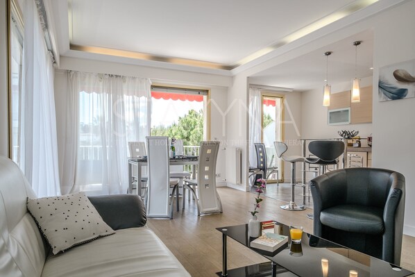A modern living room with a sofa, a coffee table, and a black leather chair. There is a dining area with a table and chairs, and a kitchen area with a counter and chairs. - Coco 2 bedroom apartment VRBO (Nice)