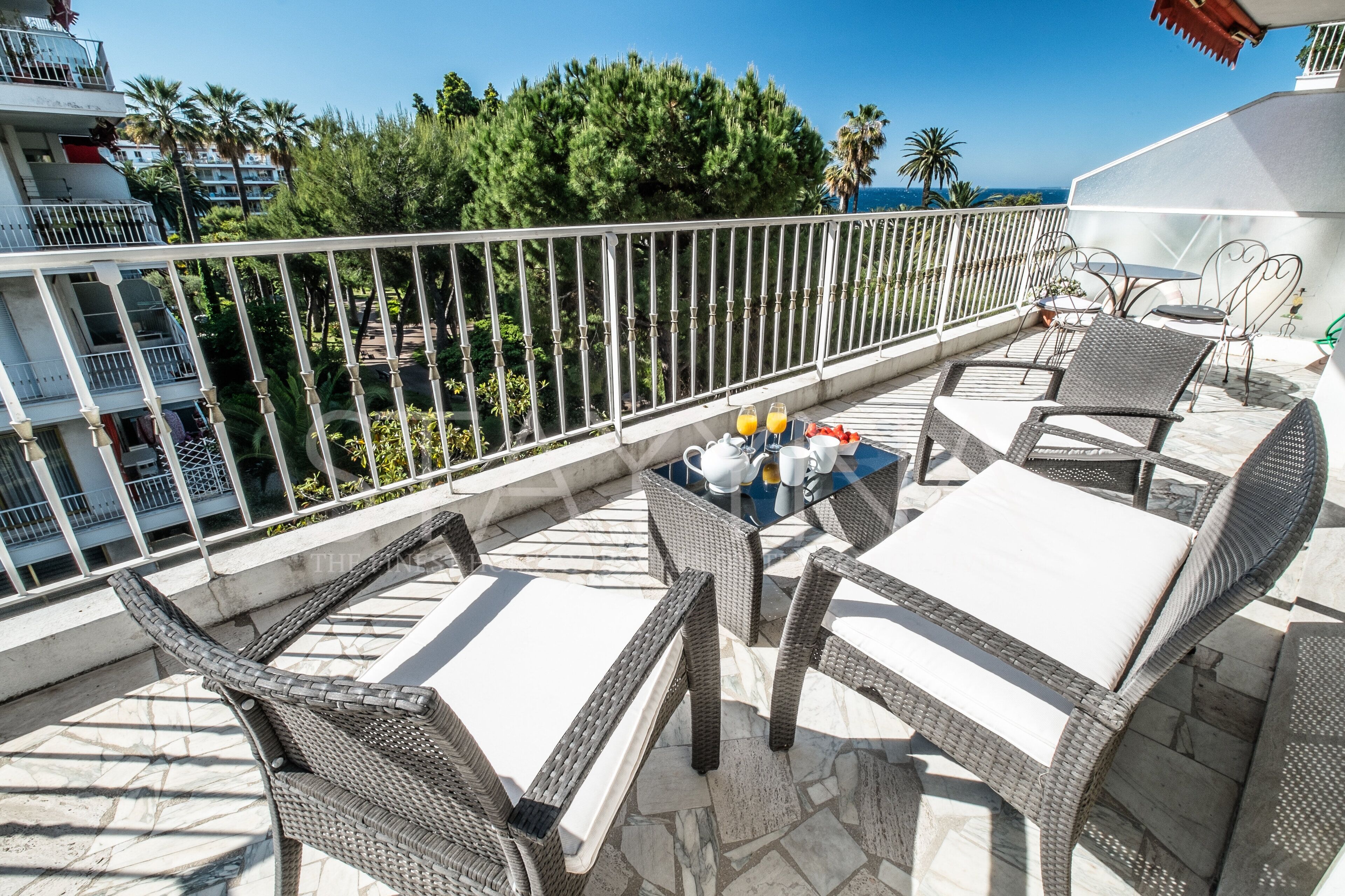 A balcony with wicker furniture, a table, and chairs, and a view of the sea and palm trees.