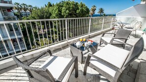 A balcony with wicker furniture, a table, and chairs, and a view of the sea and palm trees. - Coco 2 bedroom apartment VRBO (Nice)