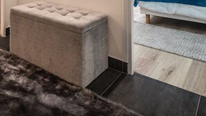 Una stanza con pavimento in legno scuro, un pouf grigio trapuntato, una panca bianca e un tappeto bianco.