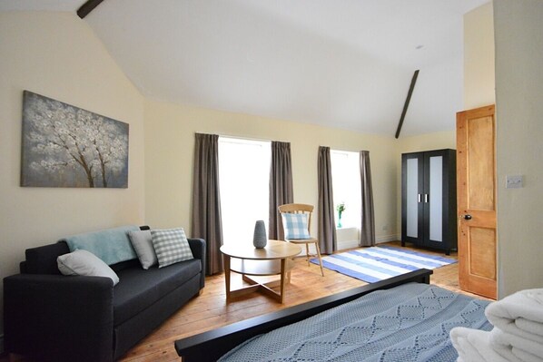 Ein modernes Zimmer mit einer schwarzen Couch, einem kleinen runden Tisch, einem Stuhl, einem Bett mit blauer Decke und einer Holztür.