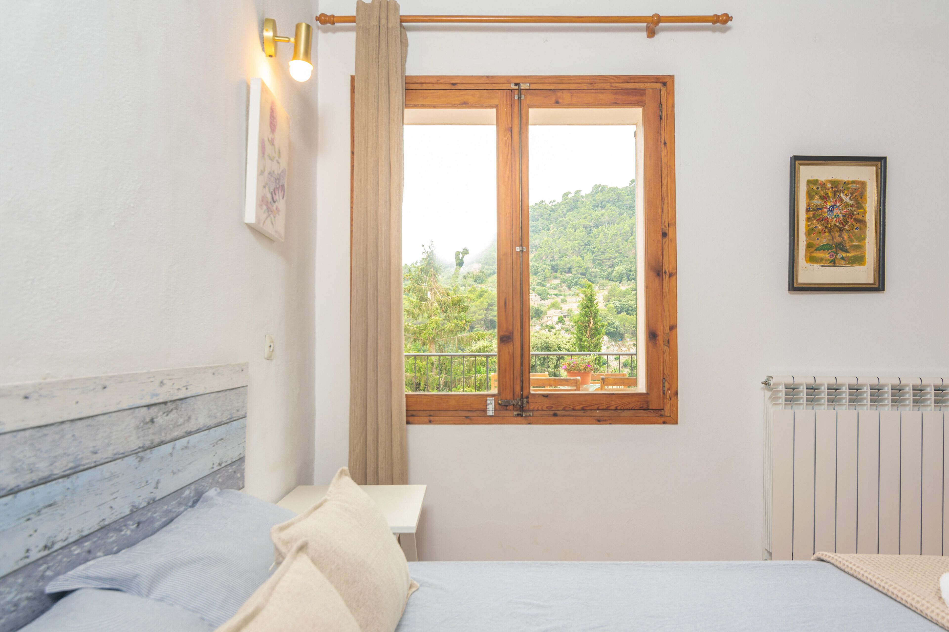 A bedroom with a bed, a window with a view of greenery, a radiator, and a wall-mounted light fixture.