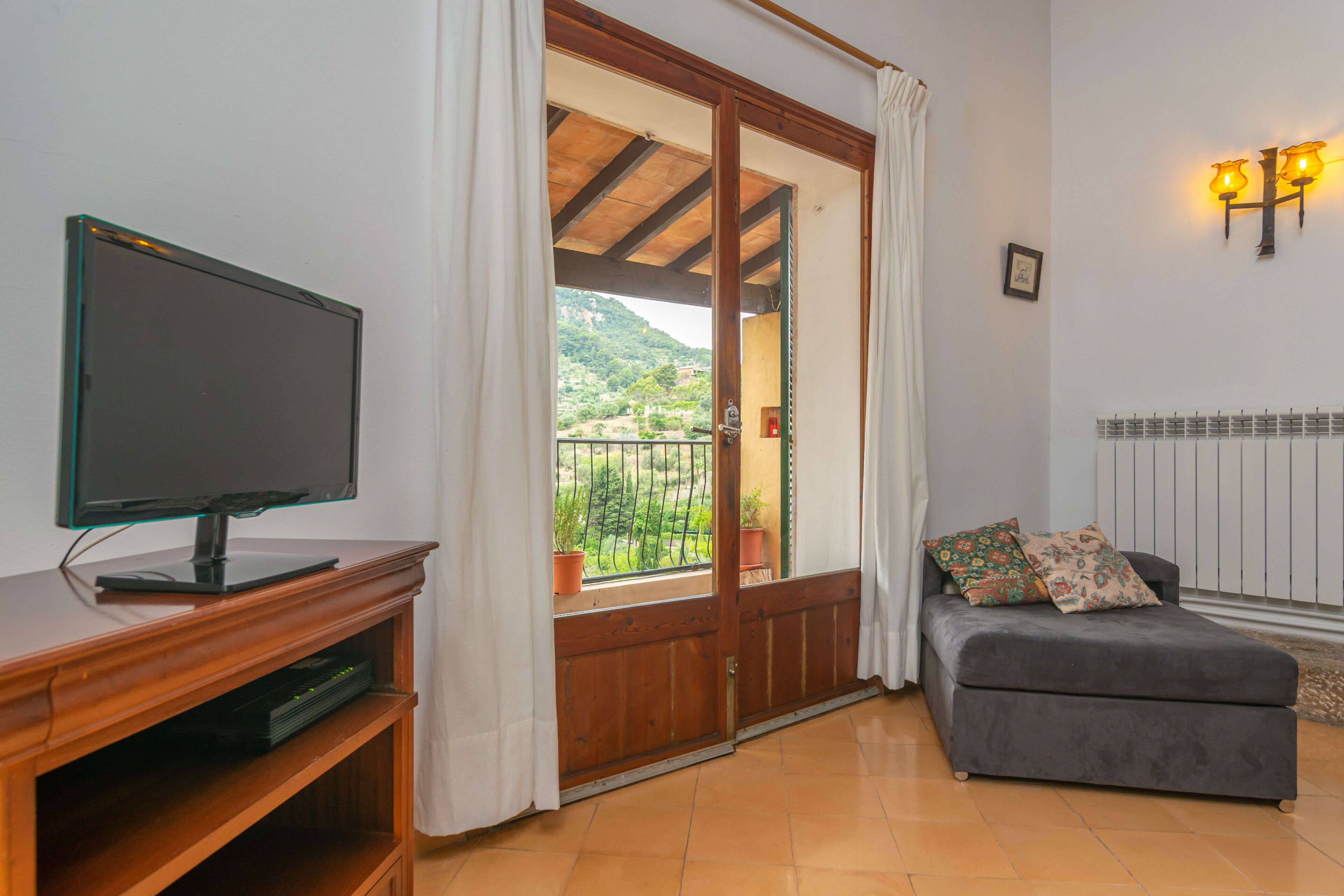 A room with a television on a wooden stand, a grey sofa with patterned cushions, a wooden door leading to a balcony, and a wall-mounted light fixture.