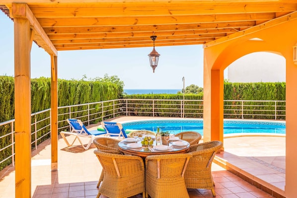 A covered outdoor dining area with wicker chairs and a table set for a meal, overlooking a swimming pool and a view of the sea.