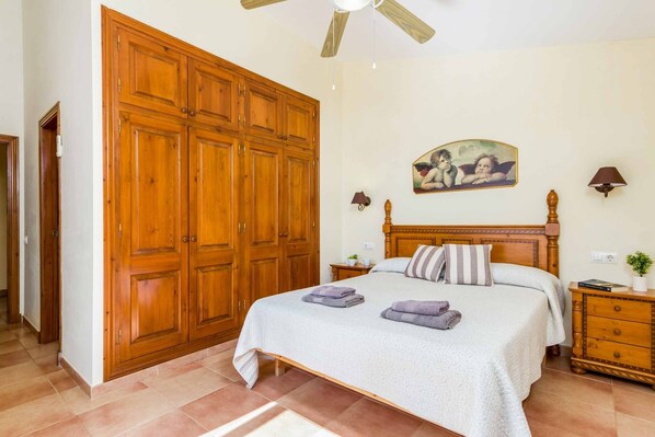 A bedroom with a large wooden wardrobe, a bed with a white bedspread, two bedside tables with lamps, and a wall-mounted painting.
