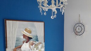 Una habitación con pared azul, sofá blanco con cojines beige, estructura de cama metálica blanca, candelabro blanco y un cuadro de una mujer junto al mar. También se observa un reloj de pared.