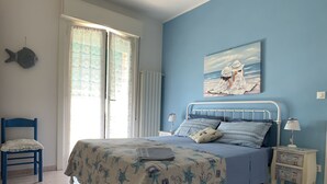 Un dormitorio con cama, una silla, una mesita de noche, una lámpara, luz en el techo, una ventana con cortinas y un cuadro en la pared.