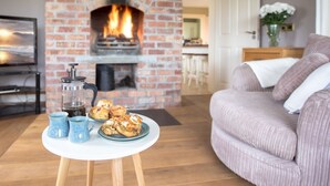 En hyggelig stue med en murstenspejs, et rundt sofabord med kager og blå krus, samt en behagelig sofa.