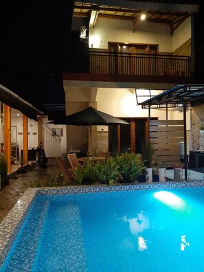 A pool area with a tiled edge, a covered seating area with chairs and an umbrella, and a balcony with wooden railings. - Indah Nusantara Homestay (Banyuwangi)