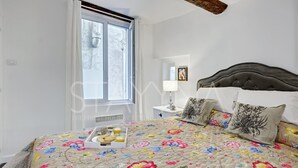 Ein Schlafzimmer mit Blumenoptik am Bett, einem Nachttisch mit Lampe und einem Fenster mit Vorhängen.