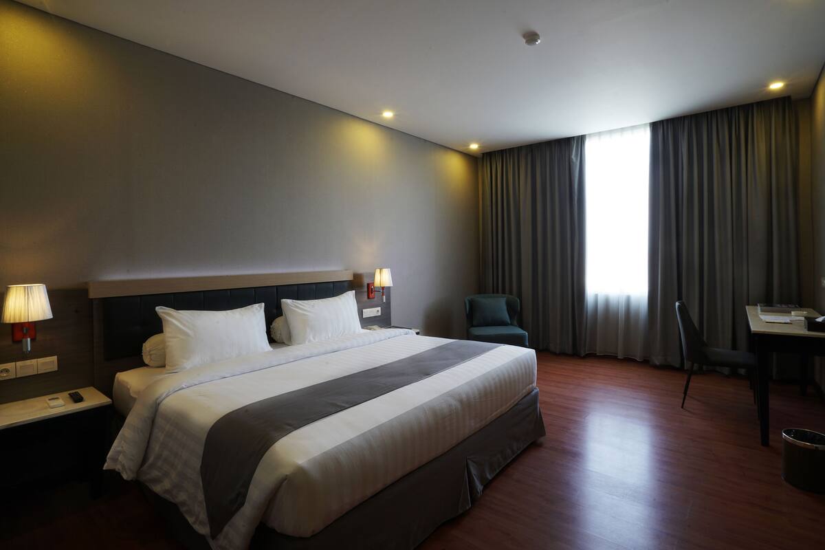 Ein Hotelzimmer mit einem großen Bett, einem Schreibtisch, einem Stuhl und einem Fenster mit Vorhängen.