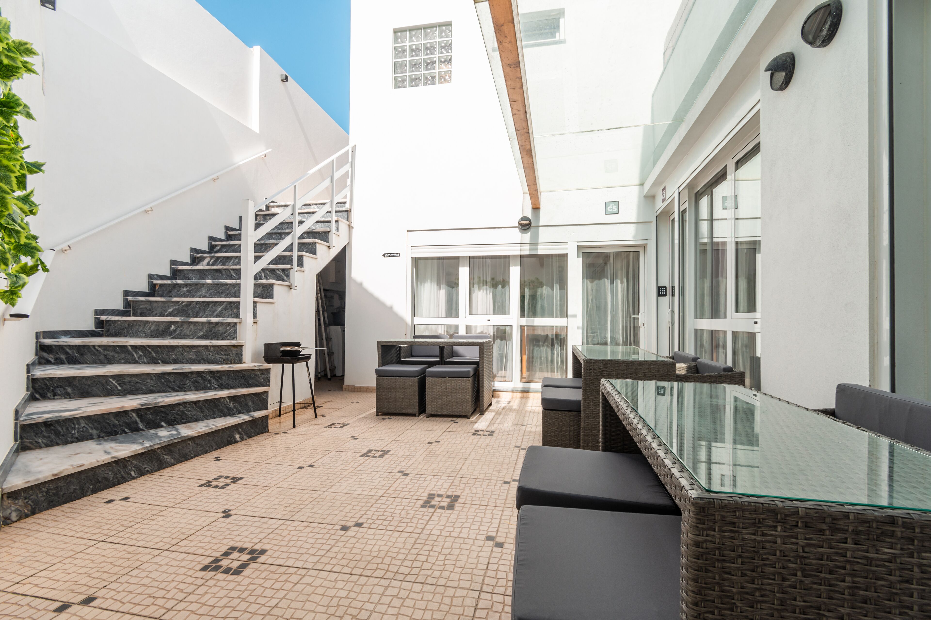 A modern outdoor patio with a glass-top table, wicker seating, and a staircase leading to an upper level.