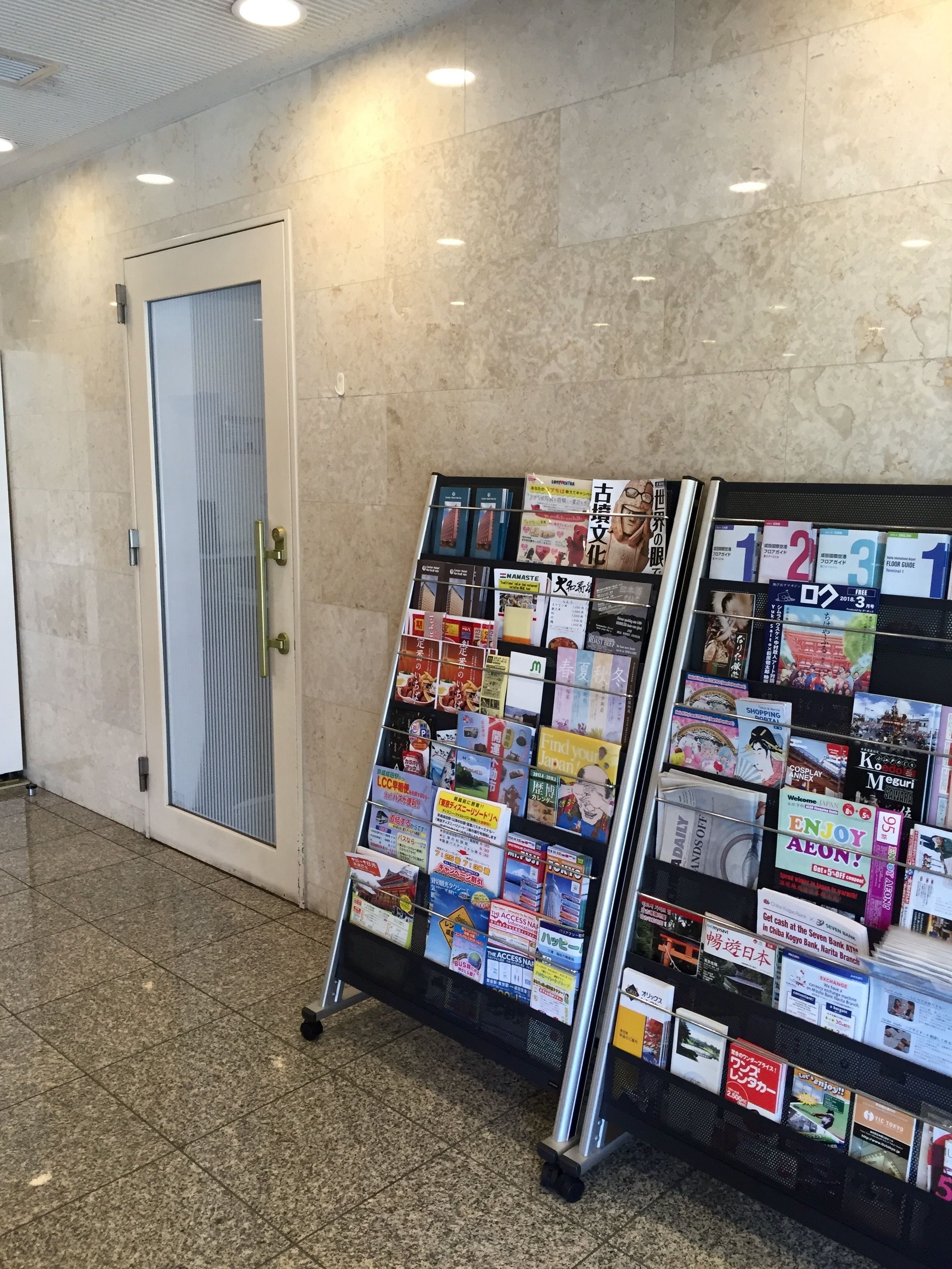 A bulletin board with various travel and informational brochures in a hallway with tiled flooring and a door.