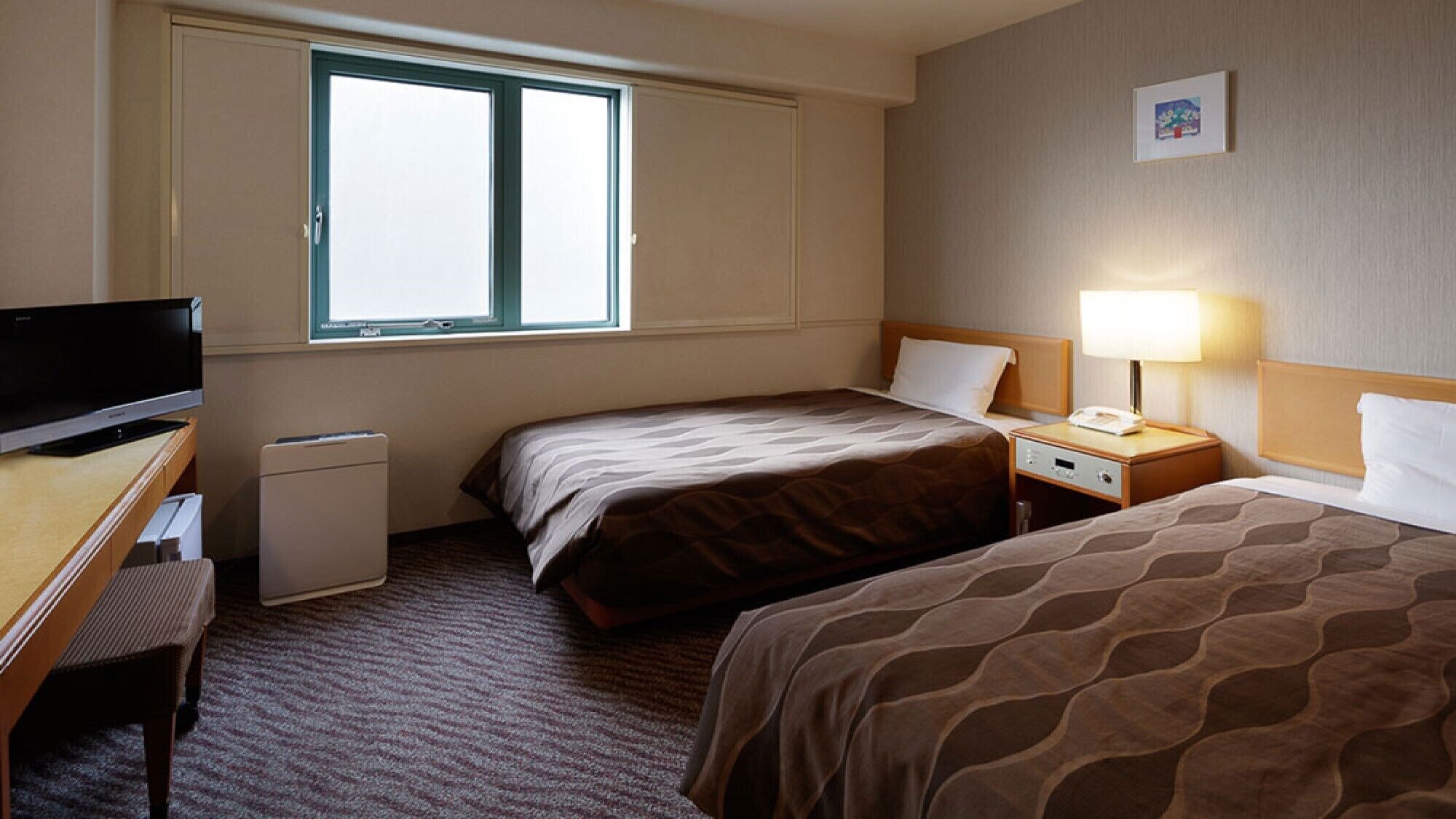 Ein Hotelzimmer mit zwei Betten, einem Schreibtisch mit Fernseher, einem kleinen Nachttisch mit Lampe und einem Fenster.