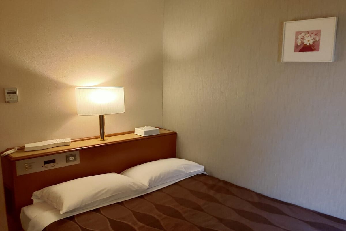 Hotelkamer met een bed, een bedlamp, een telefoon, een bedieningspaneel en een lijst in een fotokader aan de muur.