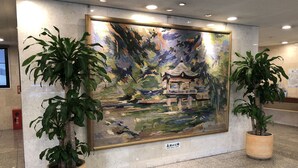 Ein gerahmtes Gemälde einer traditionellen japanischen Landschaft mit einem Pavillon, das von zwei Topfpflanzen gesäumt wird, auf einem Fliesenboden.