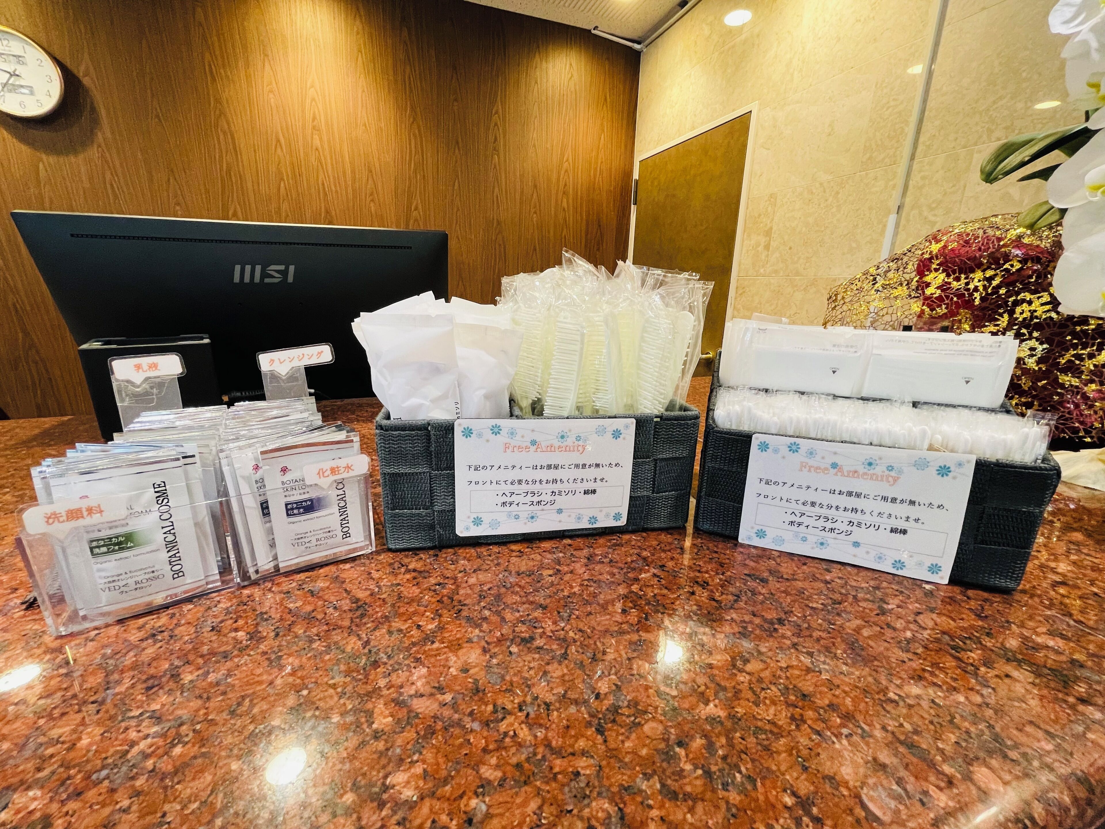 A hotel reception area with a wooden panel wall, a reception desk, and a computer monitor. There are signs indicating amenities such as free amenities, and various toiletries and tissues are neatly arranged on the counter.