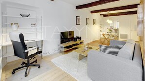 A modern living room with a white desk, a black office chair, a grey sofa, and a wooden coffee table. There is a TV on a stand, a bookshelf, and a dining area in the background. - La Caravelle 1 bedroom apartment VRBO (Nice)