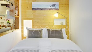 A neatly made bed with a white comforter and pillows, a wooden headboard, a bedside lamp, and a framed picture of a beach scene. - La Caravelle 1 bedroom apartment VRBO (Nice)