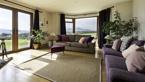 A spacious living room with a large window offering a view of the countryside. There is a sofa set, a coffee table, and a potted plant. - The Lookout 3 bedroom house (Dingle Peninsula)