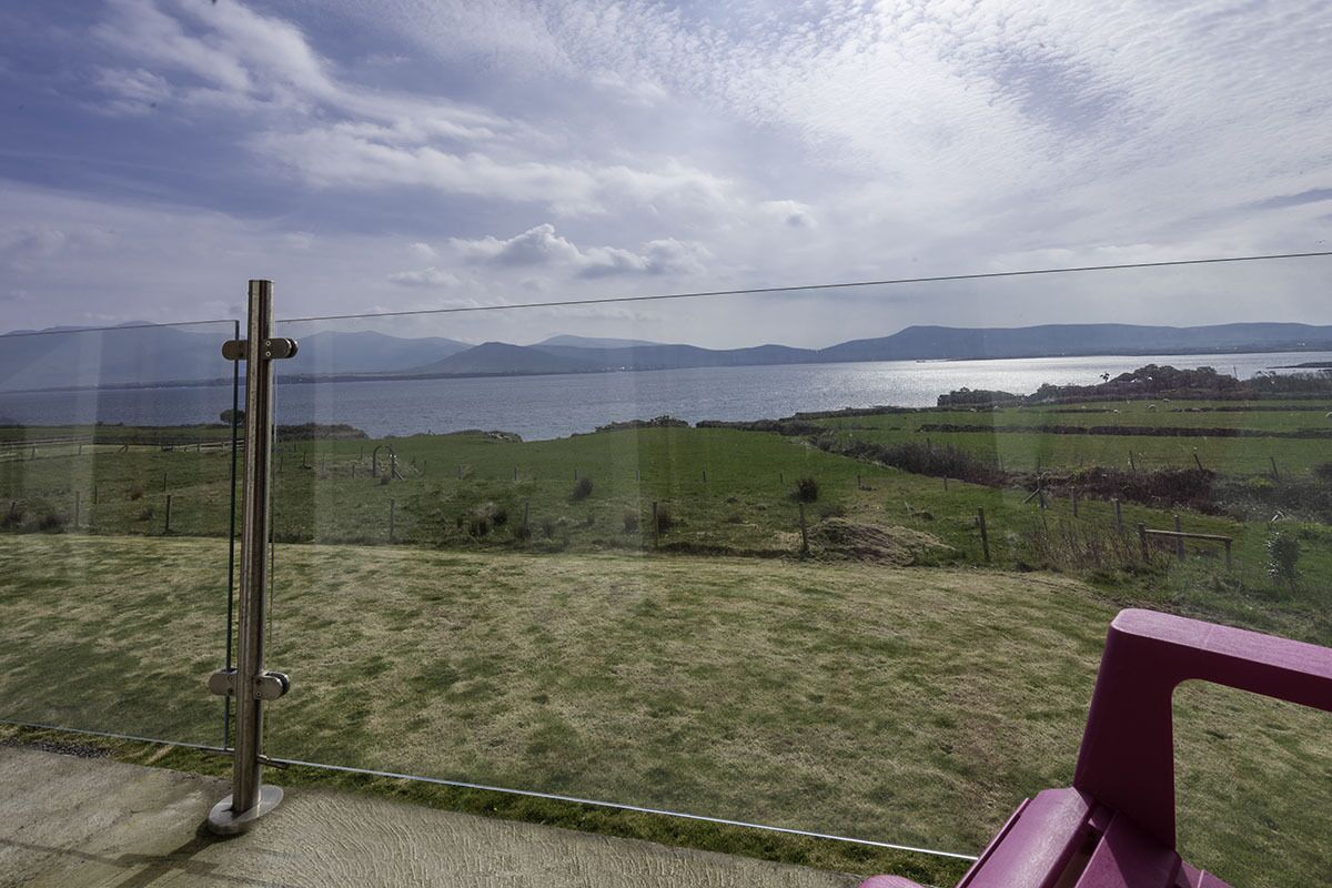 A view from a balcony overlooking a body of water with distant mountains, a grassy field, and a metal structure.