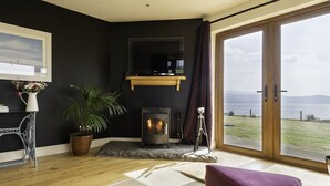 A cozy room with a fireplace, a large window with a view of the sea, and a wooden floor. - The Lookout 3 bedroom house (Dingle Peninsula)