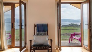 A room with a wooden chair and a blue blanket, large glass doors leading to a balcony with a pink chair and a view of the countryside. - The Lookout 3 bedroom house (Dingle Peninsula)