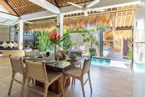 Un espace repas avec vue sur la piscine, comprenant une table dressée pour quatre personnes, entourée de chaises en osier, et un jardin tropical doté d’un toit de chaume.