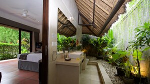 Habitación con cama, lavamanos y zona de ducha. Hay plantas y techo de paja.