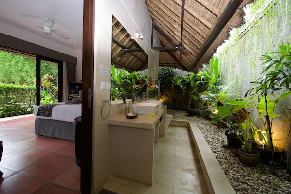 Habitación con cama, lavamanos y zona de ducha. Hay plantas y techo de paja.