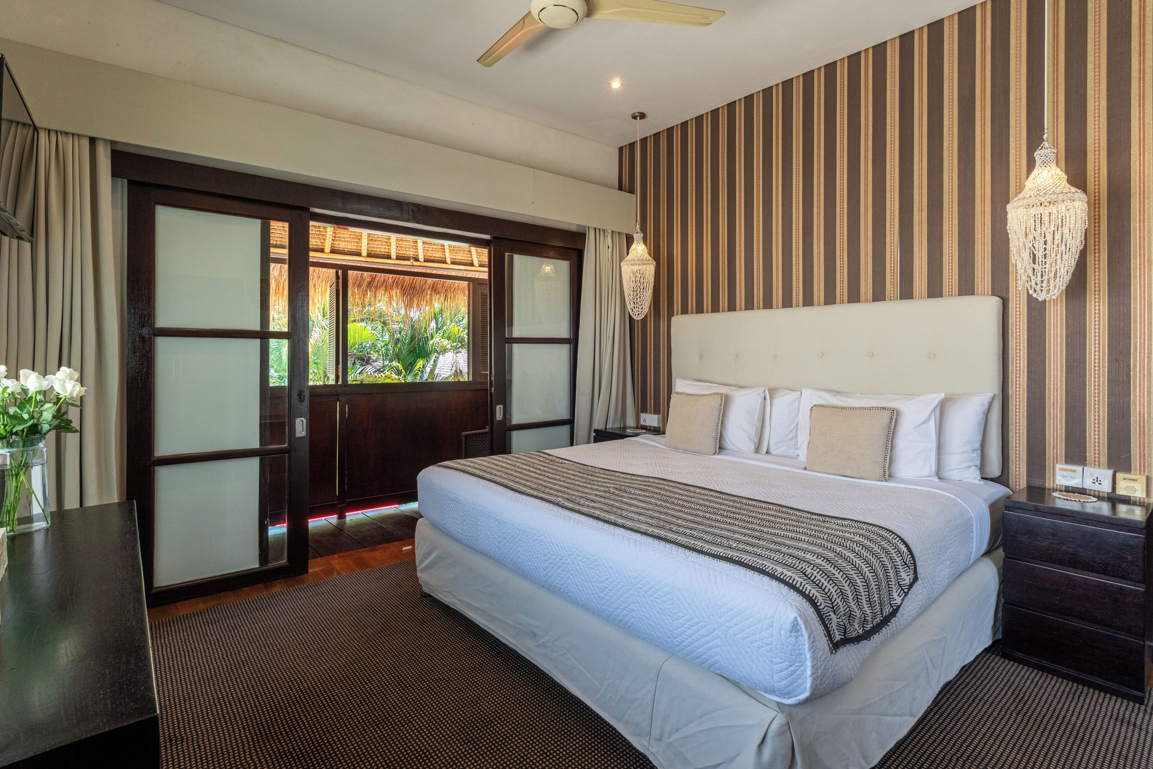 A hotel room with a large bed, a ceiling fan, a view through a glass door, and a striped wall.