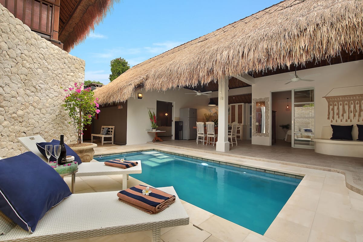 A pool area with lounge chairs, a thatched roof, and a stone wall.