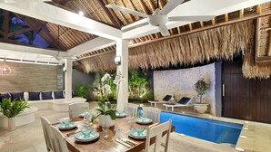 Un área de comedor junto a la piscina con una mesa de madera dispuesta para cuatro personas, rodeada de sillas de mimbre blanco.